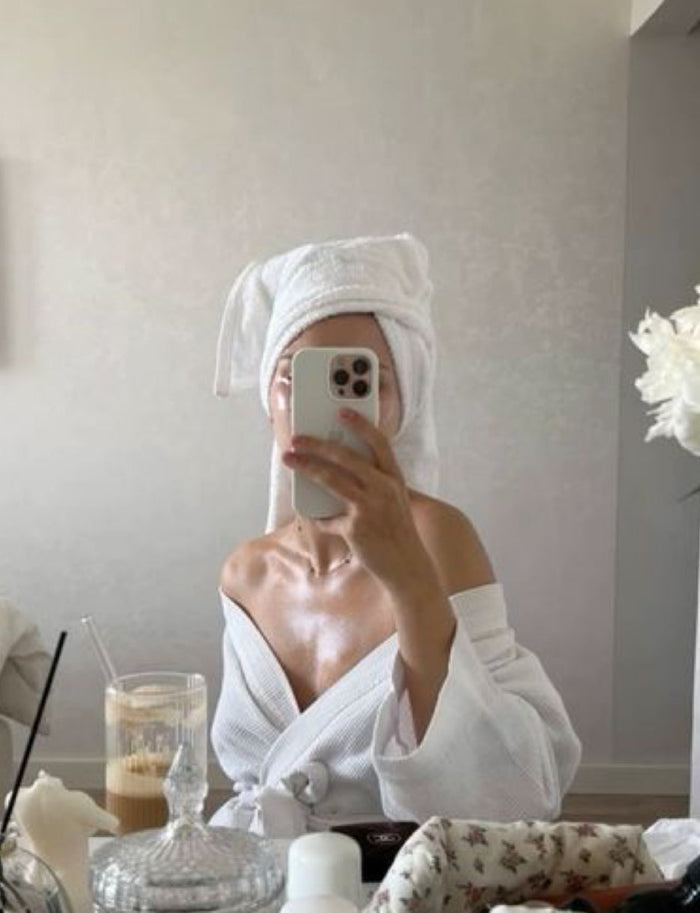 Model wearing a white robe and towel wrapped around her head, taking a picture in the mirror 