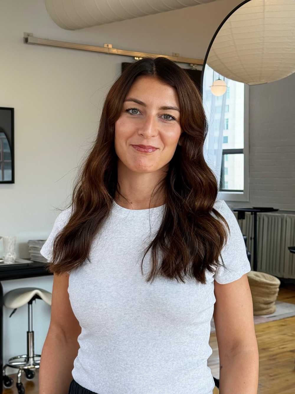 Priscilla Sorin hairstylist at Private Studio hair salon in Toronto's Fashion District. A woman with long brown hair wearing a light gray short-sleeve crewneck t-shirt, standing in a modern office or studio environment with a window, radiator, and hanging paper lantern visible in the background.